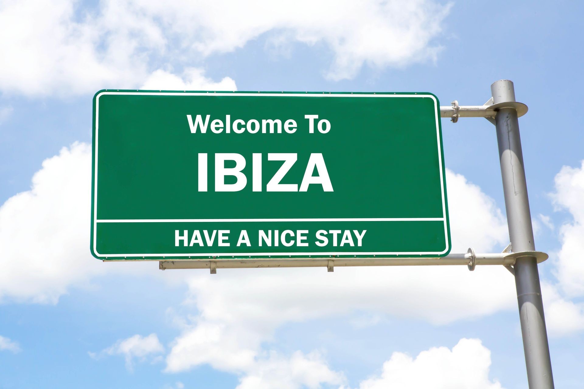 Green overhead road sign with a Welcome to Ibiza, Have a Nice Stay concept against a partly cloudy sky background.